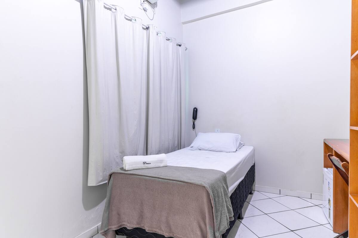 A small, clean room with a bed, a bedside table, a wall-mounted phone, and a wooden shelf.