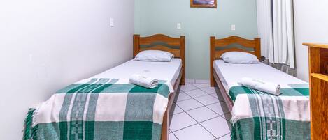 Two single beds with wooden frames and green and white checkered blankets, a small framed picture on the wall, and a white curtain on the right.