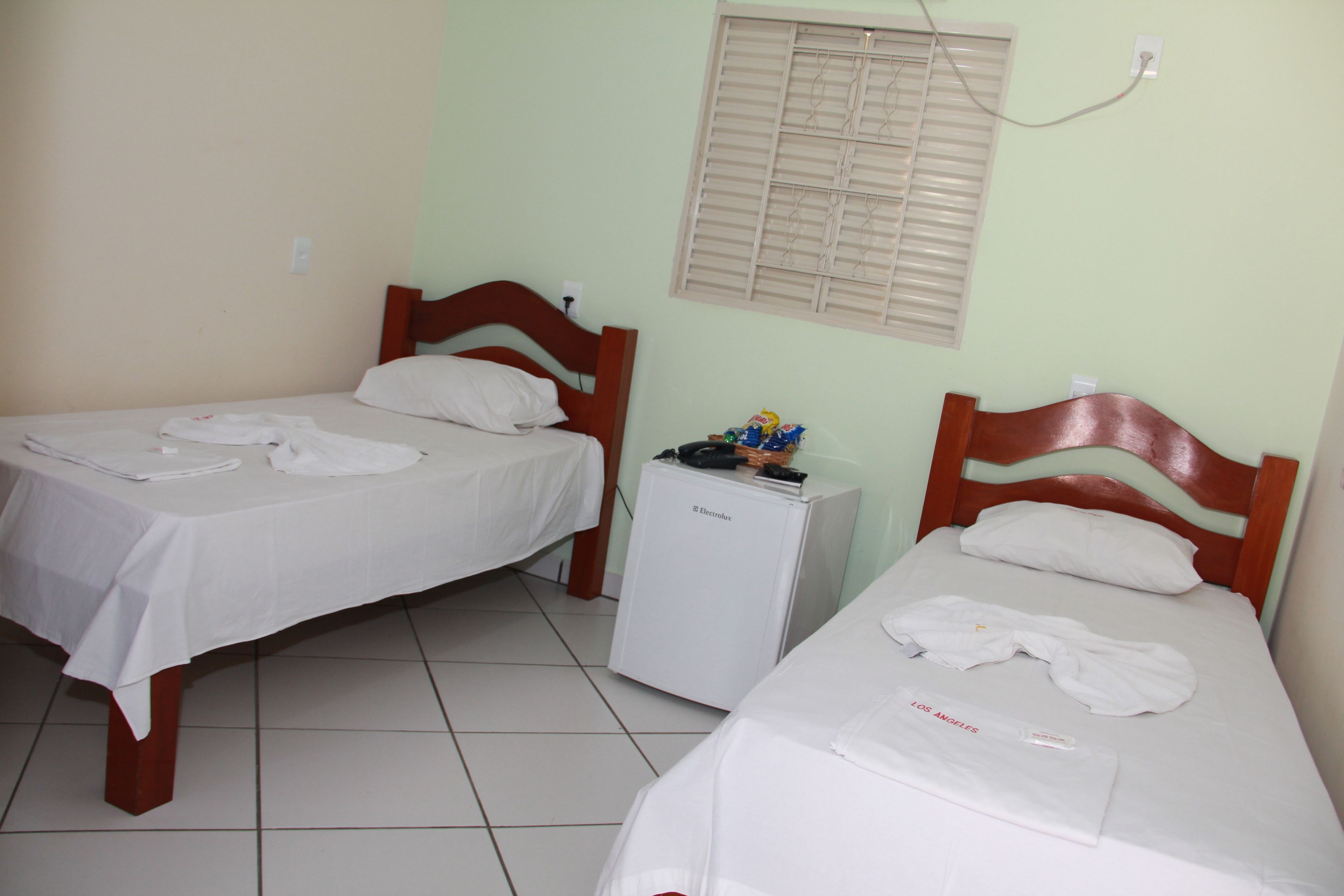 Quarto pequeno e limpo com duas camas de solteiro, um pequeno frigorífico e janela com persianas.