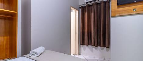 A hotel room with a bed, a wooden wardrobe, a television mounted on the wall, and an air conditioning unit.