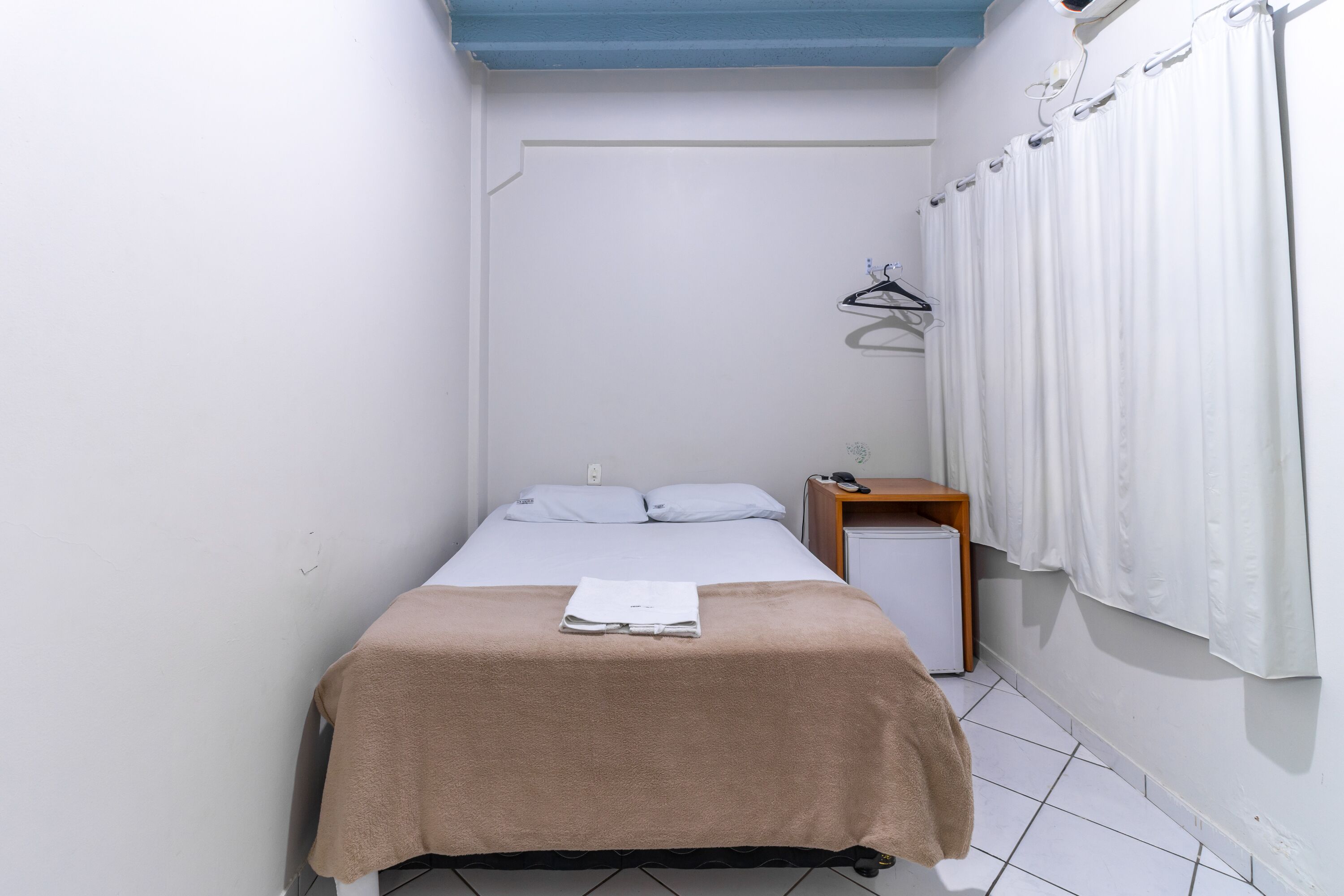 Ein kleines Zimmer mit einem Einzelbett, einer beigen Bettdecke, weißen Kissen und einem Nachttisch mit Lampe.