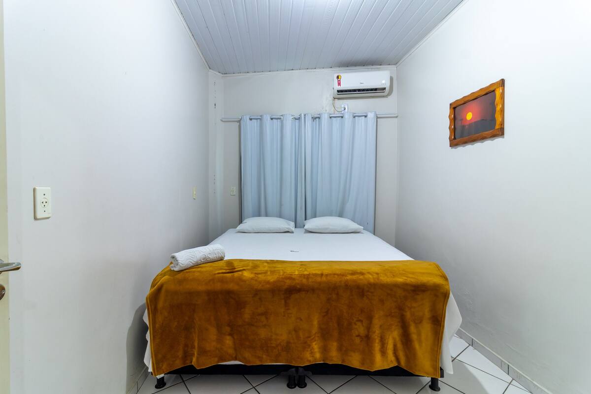 A small, single-bed room with a white ceiling, a wall-mounted air conditioner, a curtain, and a framed picture.