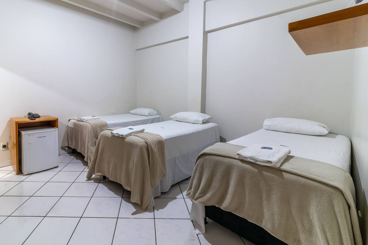 A hotel room with three single beds, each with a white mattress and a beige blanket. There is a small wooden nightstand with a phone and a white cabinet.