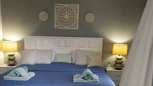 Un letto rifatto con cura, con una coperta blu, una testiera bianca e numerosi cuscini. Due abat-jour con paralumi, un elemento decorativo sulla parete sopra il letto e una tenda parzialmente aperta.