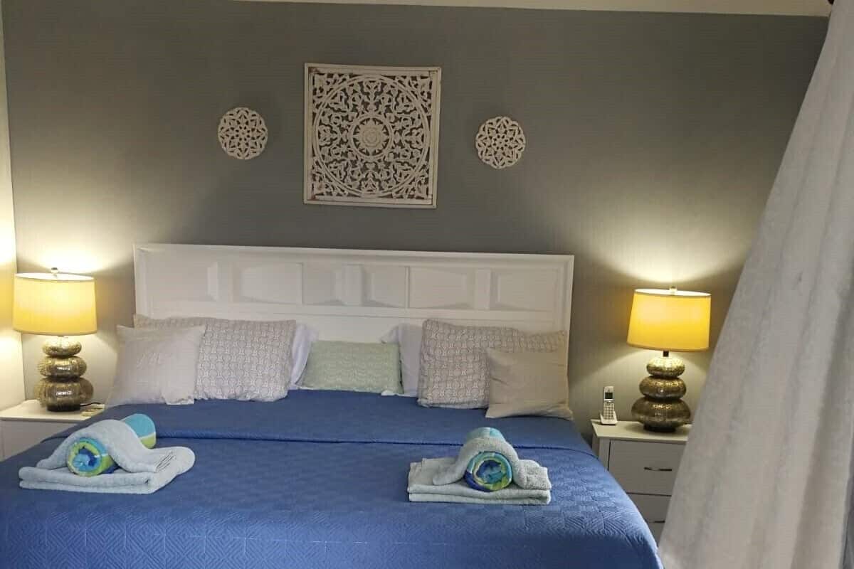 Un lit bien fait, avec une couvre-lit bleue, une tête de lit blanche et plusieurs oreillers. On y trouve deux lampes de chevet avec des abat-jour, un élément décoratif accroché au mur au-dessus du lit, et un rideau partiellement tiré sur le côté.