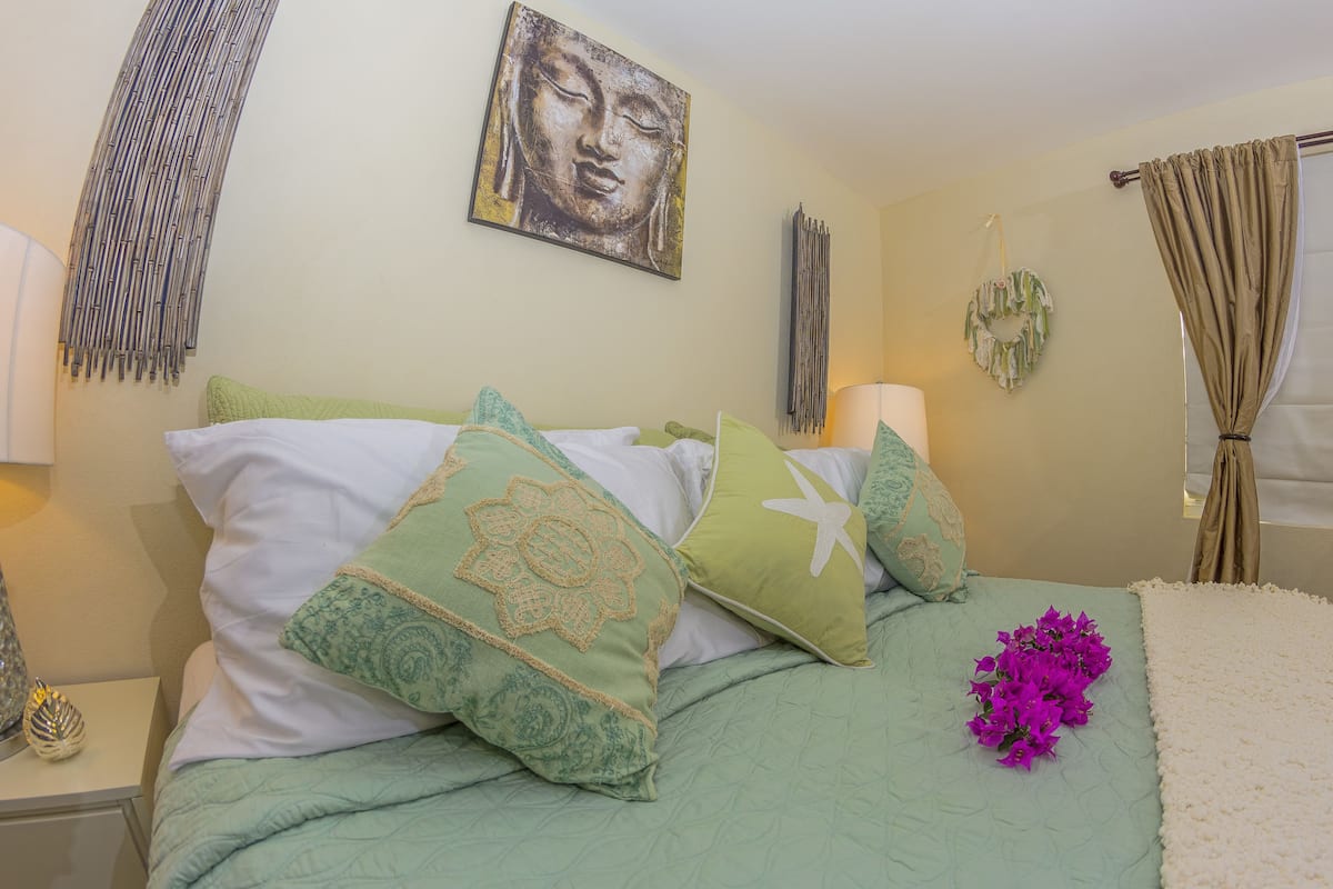 Une chambre à coucher avec une couvre-lit verte, des coussins décoratifs, une œuvre d’art accrochée au mur et une fenêtre avec des rideaux.