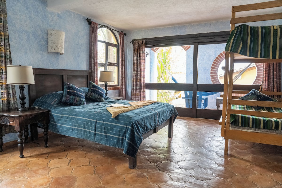 Ein Zimmer mit einem Bett, einer Holzbank, einem Tisch mit Lampe und Blick ins Freie.