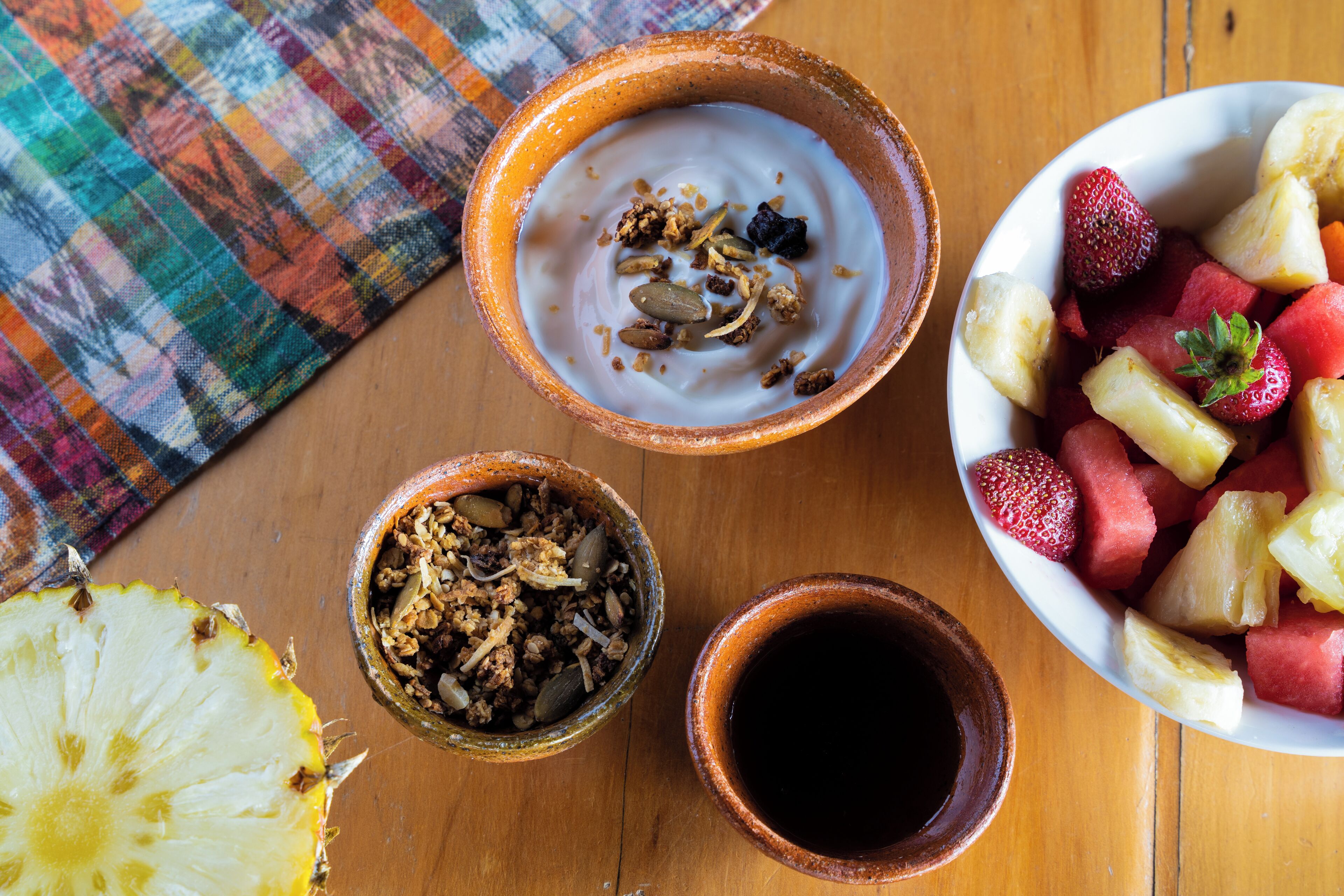 A wooden table with bowls of yogurt, granola, and fruit, and a slice of pineapple.