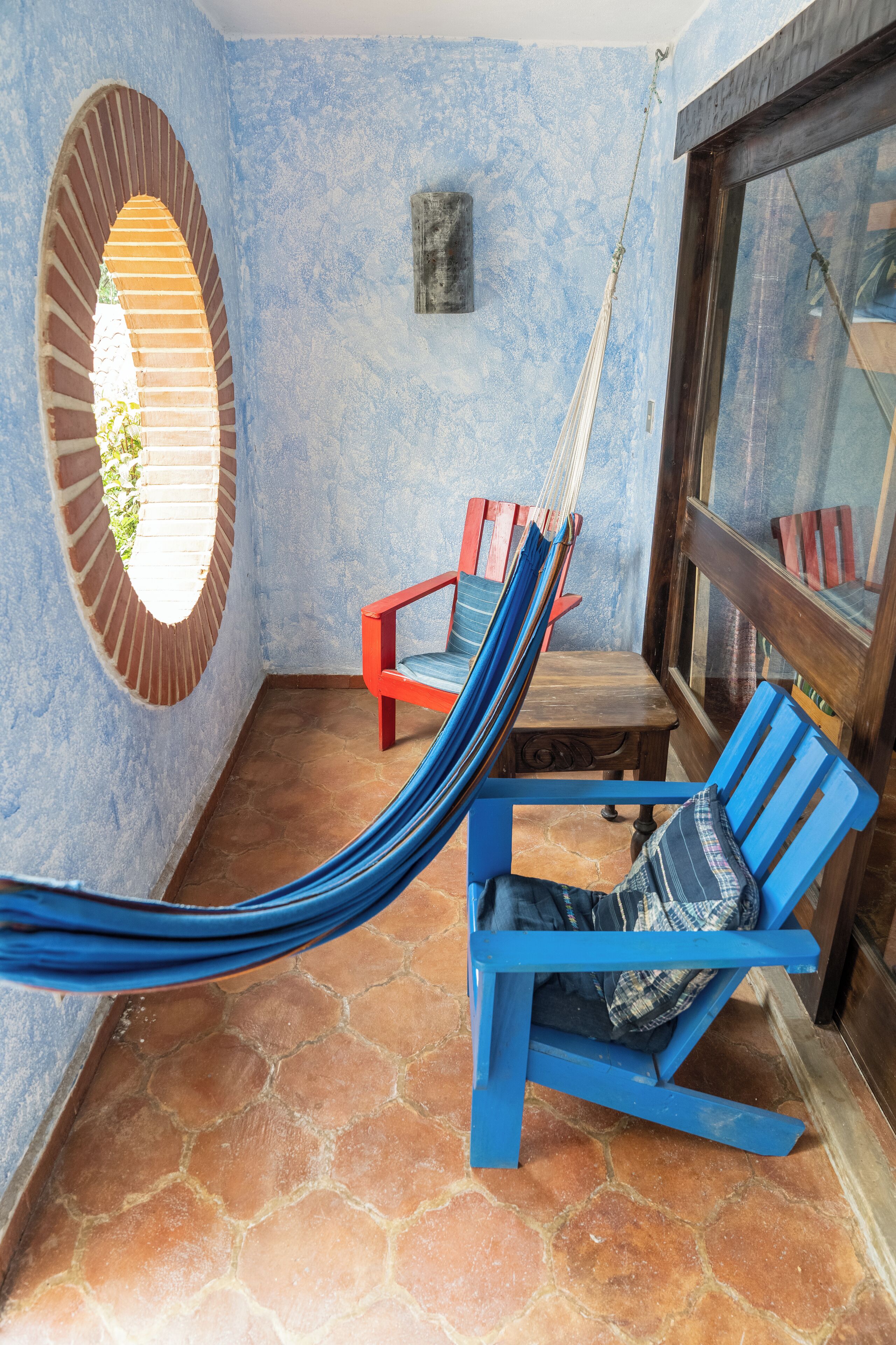 Un hamac tendu entre deux chaises bleues, posées sur un sol carrelé, avec une arche en briques et une fenêtre en arrière-plan.