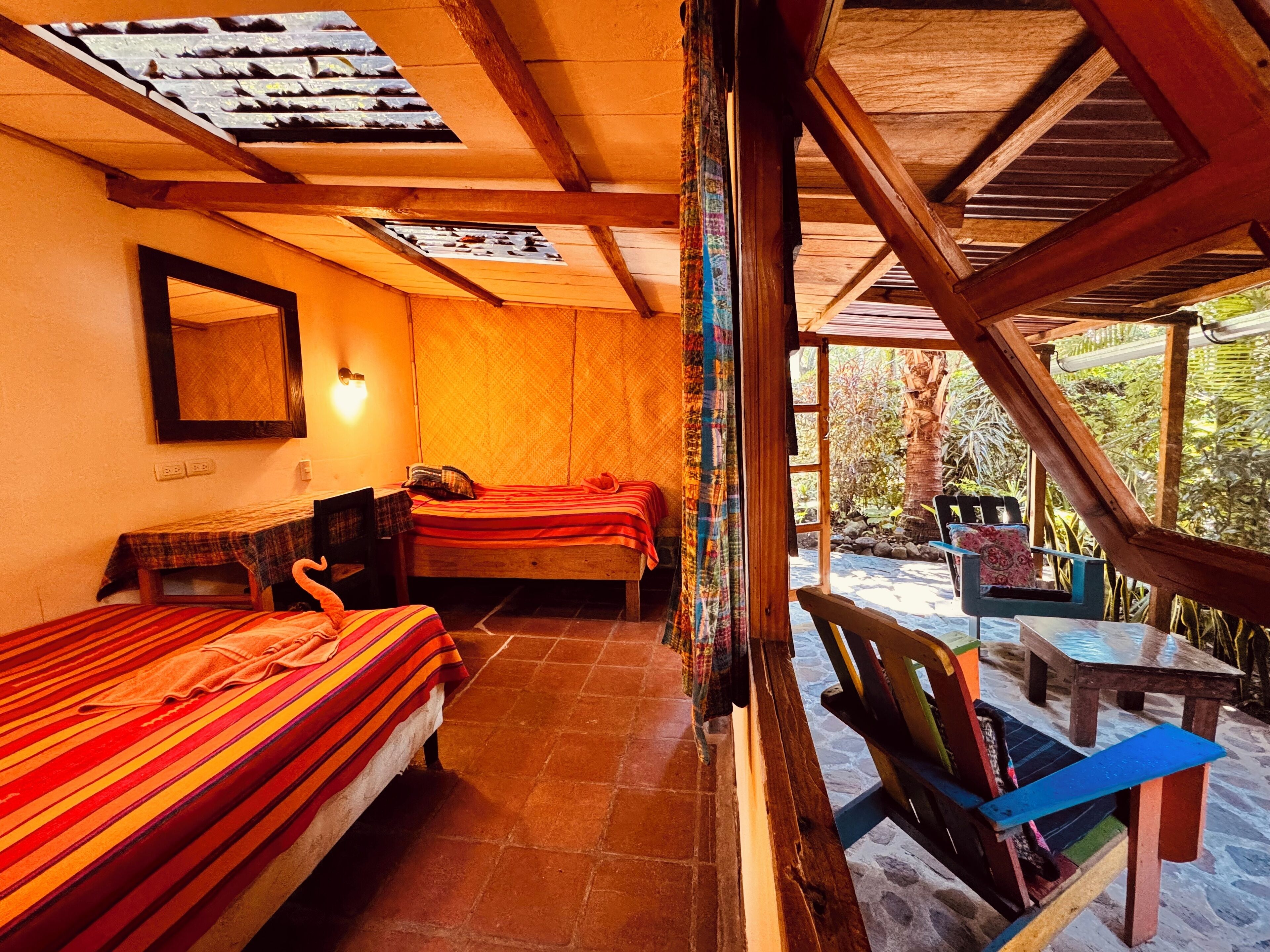 Una habitación acogedora con dos camas, una mesita y un banco. Se ve vegetación a través de la ventana.