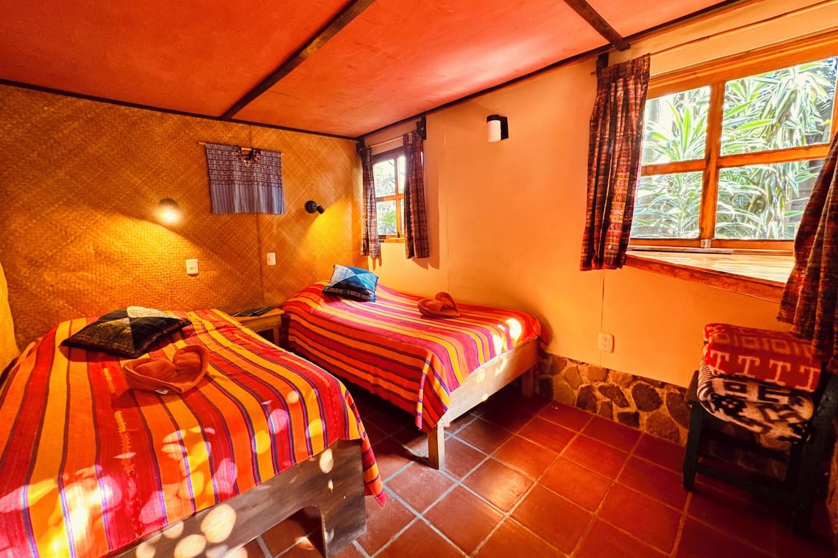 Ein Zimmer mit zwei Betten, einem Vorhangfenster, einer Wandlampe und einem kleinen Tisch.