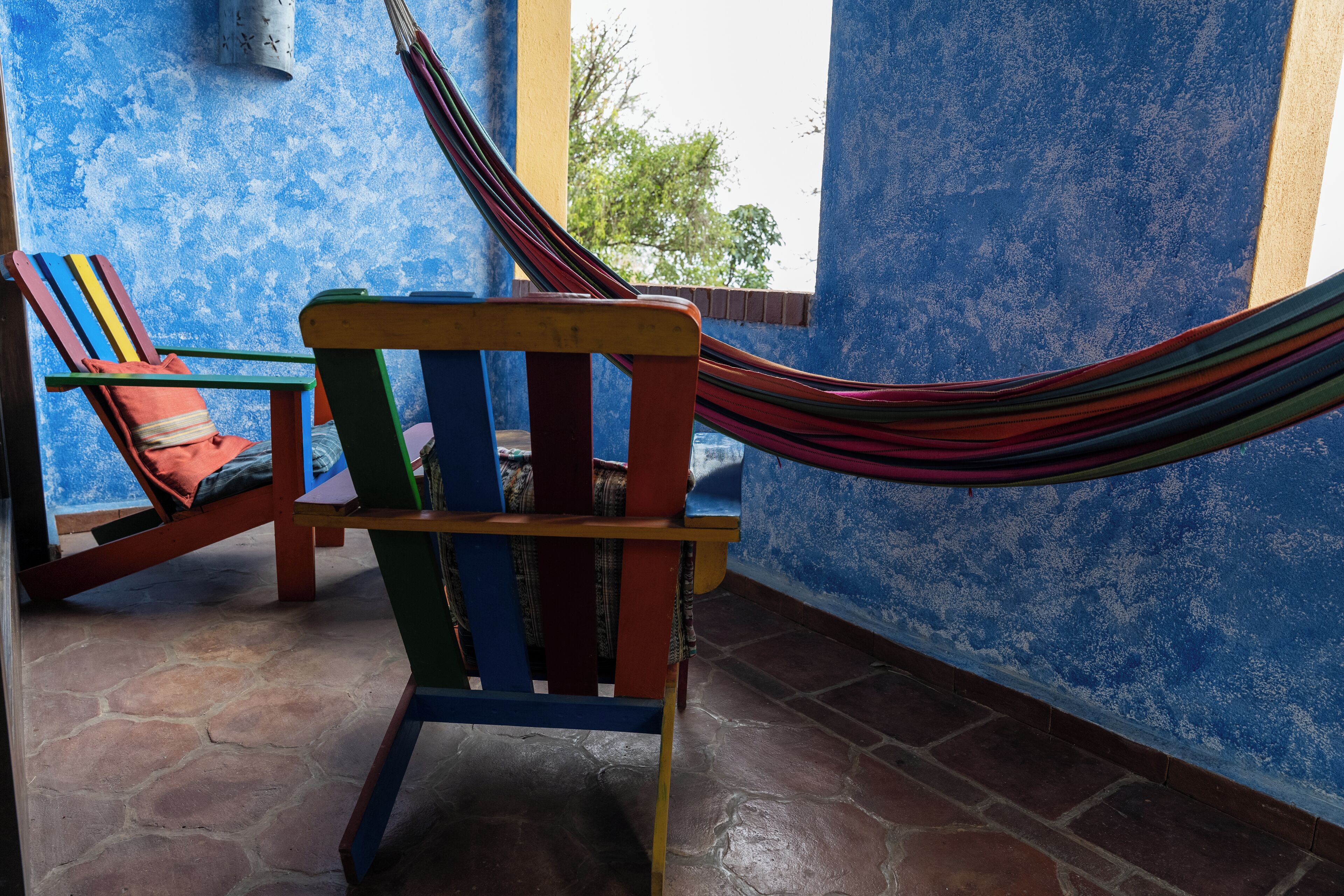 Una silla hamaca con una hamaca a rayas, una silla de madera con cojín y una pared azul con textura.