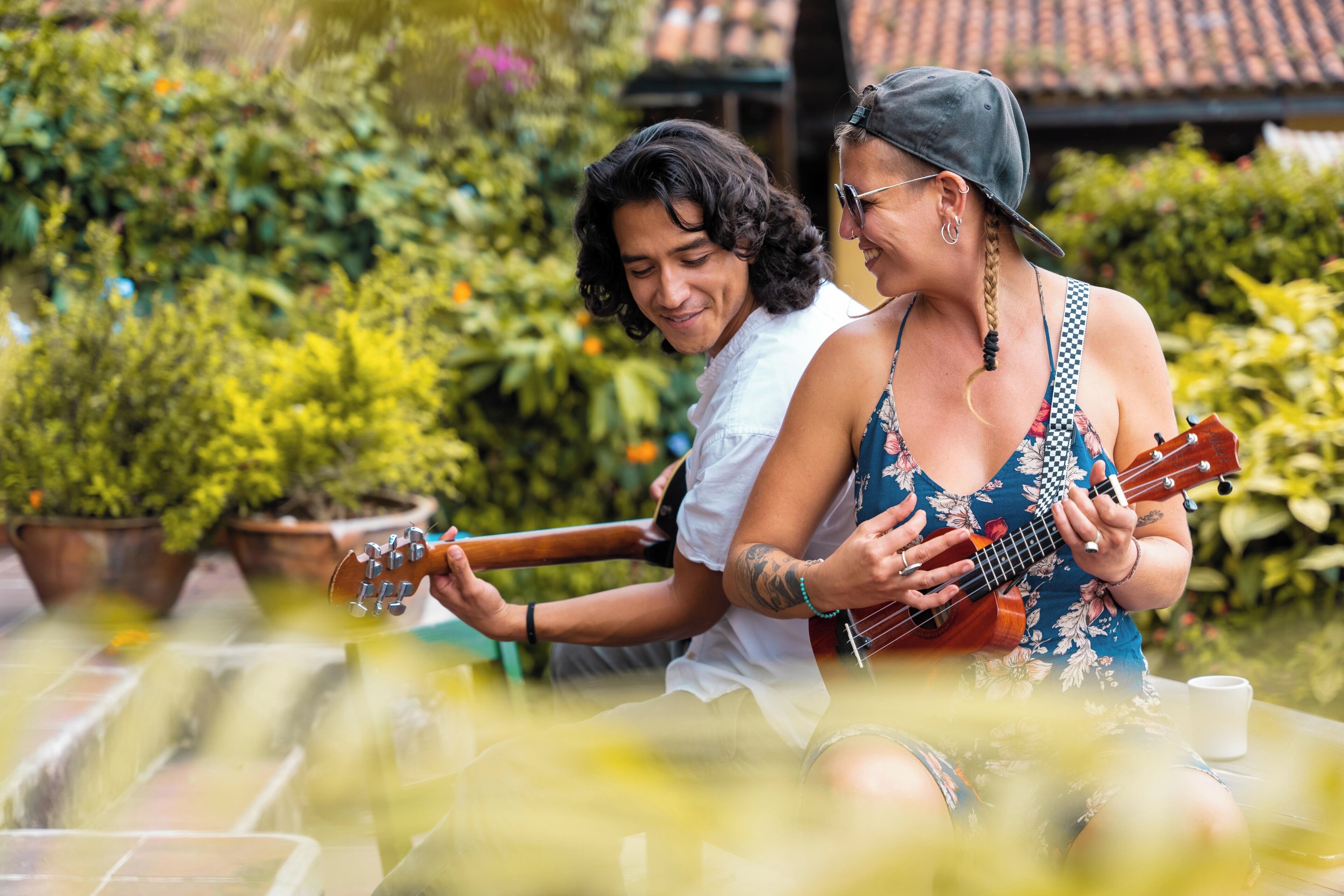 Dos personas tocan ukeleles al aire libre, con macetas y un tejado de baldosas al fondo.