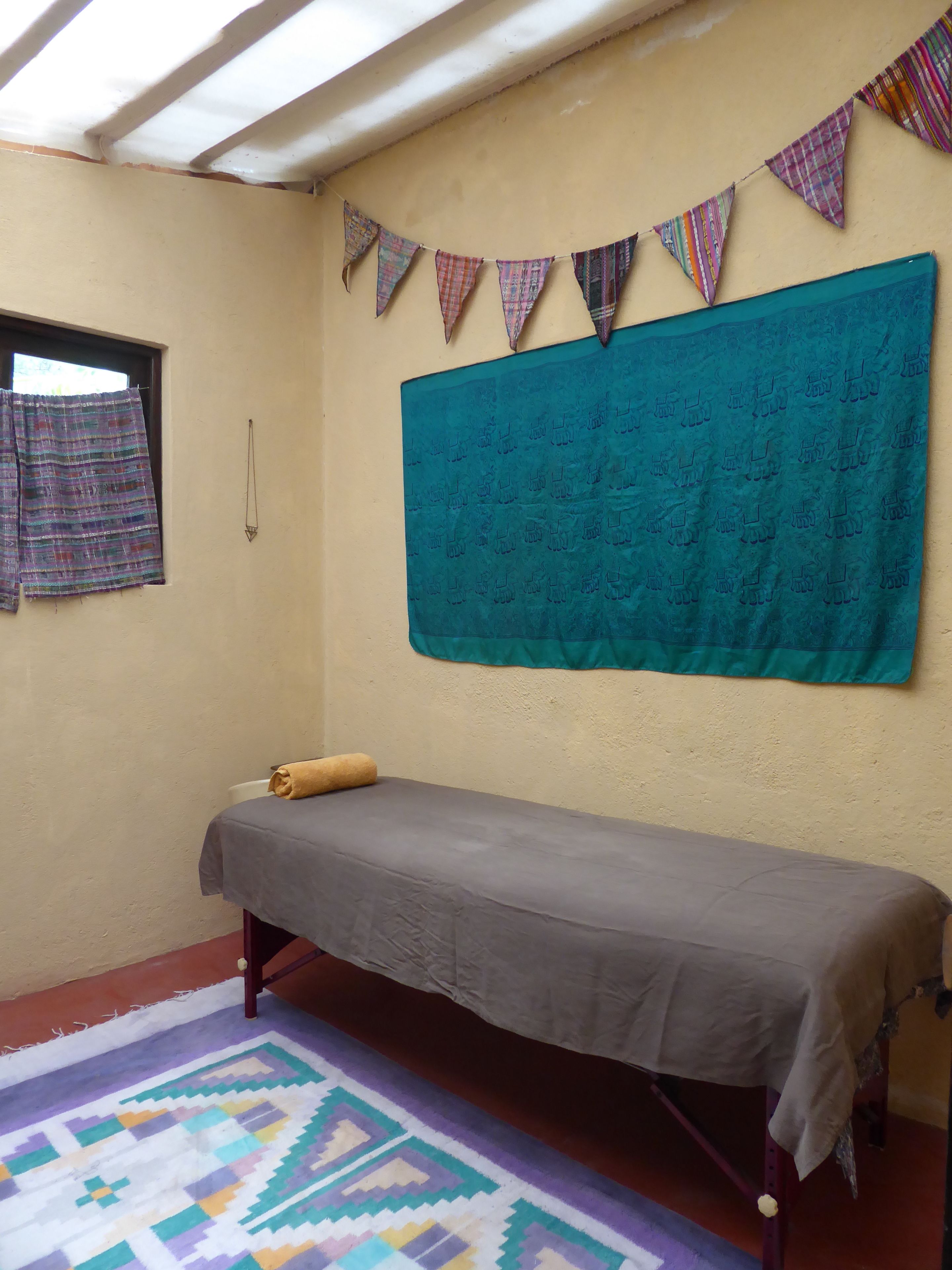 A small room with a single bed, a window with a curtain, a wall hanging, and a patterned rug.