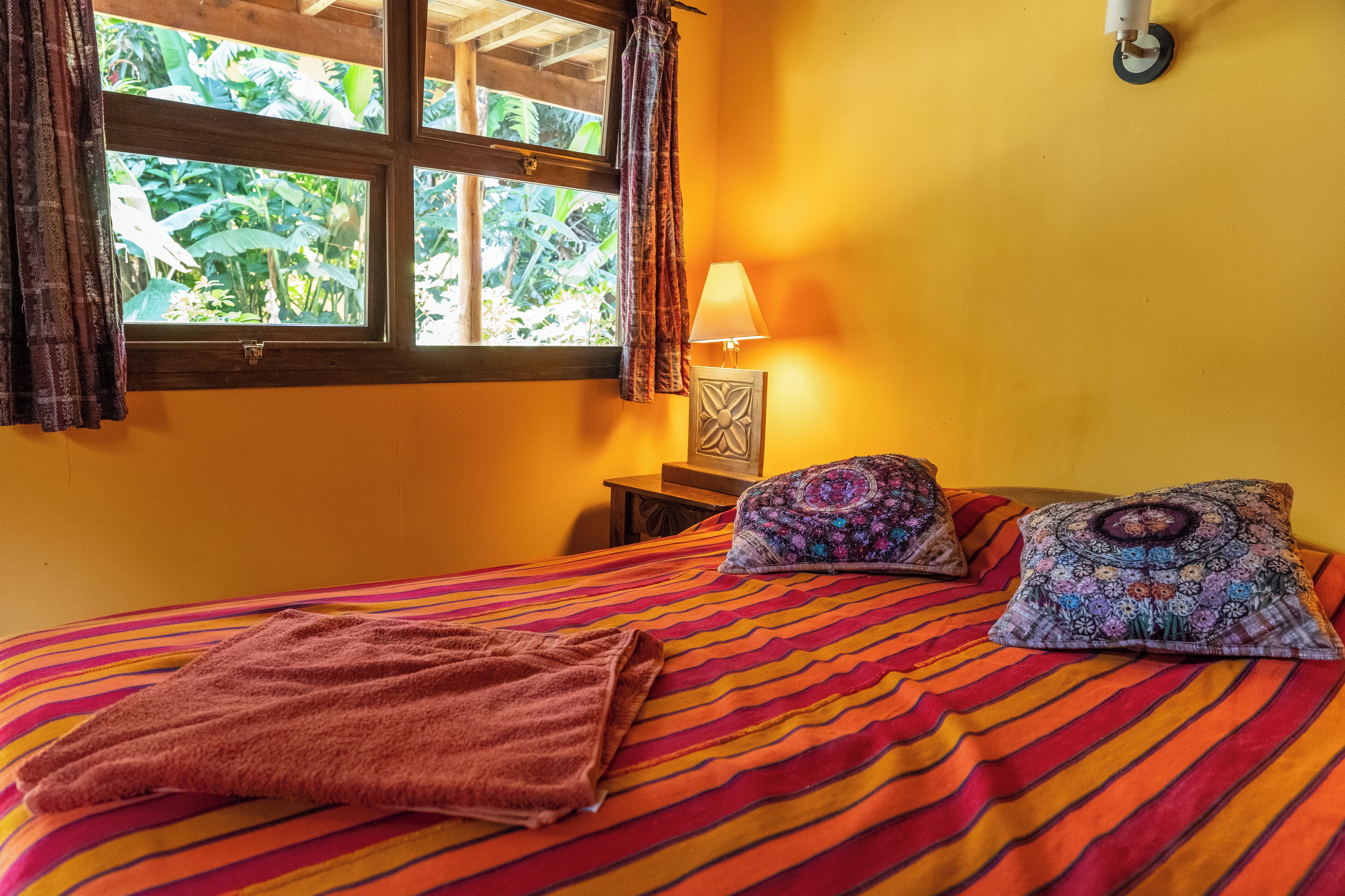 Una cama con colcha a rayas, dos almohadas estampadas, una lámpara de noche y una ventana con cortinas.