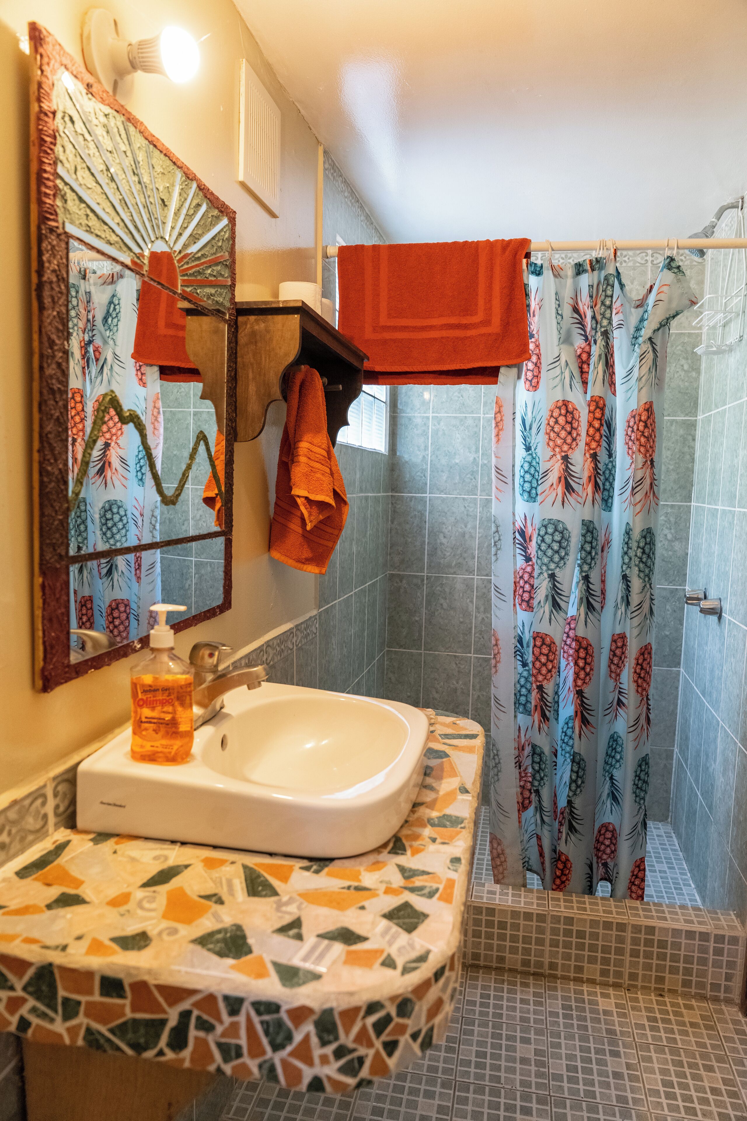 Un bagno con un lavandino bianco, uno specchio, una tenda da doccia con motivo di ananas e un pavimento piastrellato.