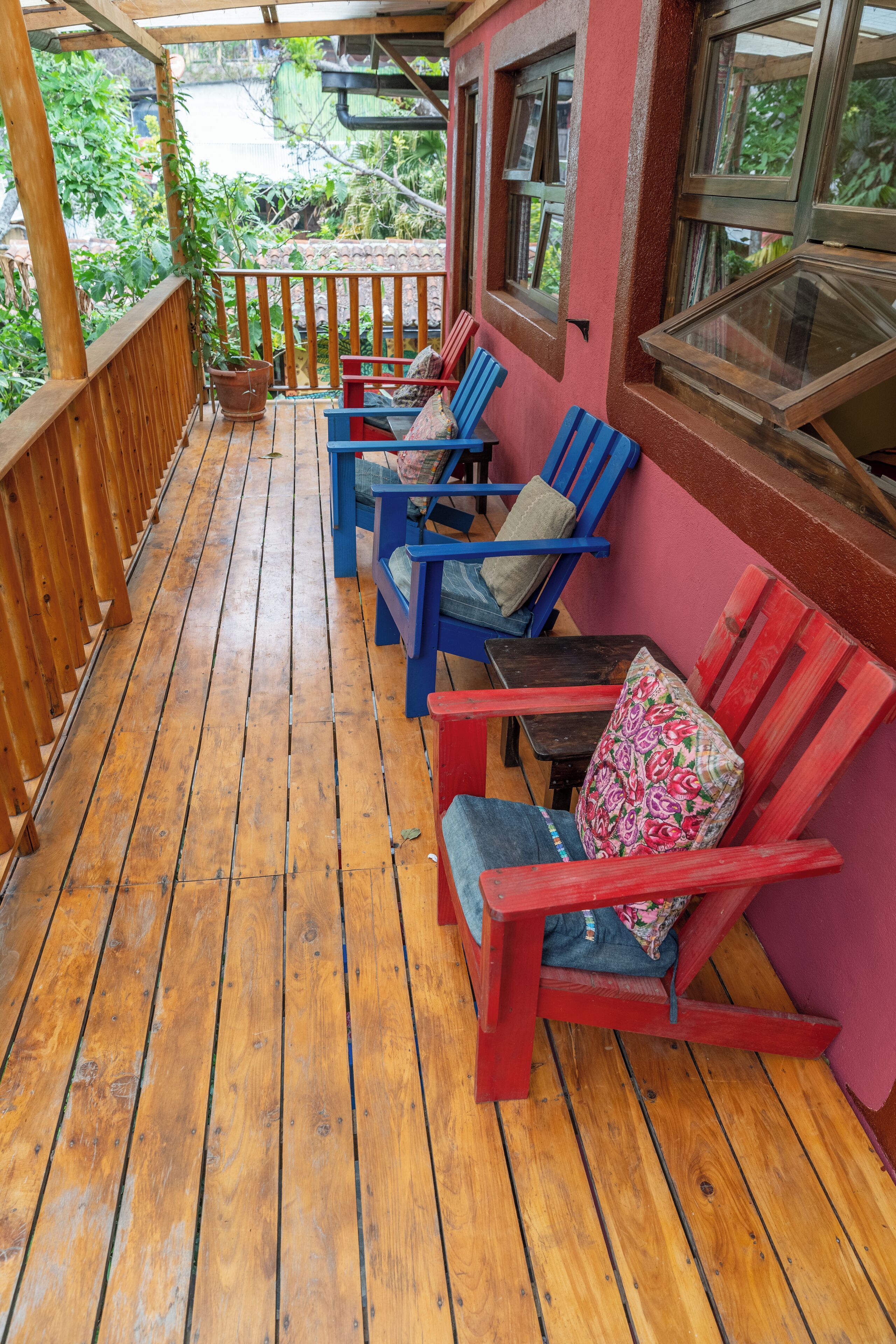 A wooden deck with red and blue chairs, a wooden railing, and a view of greenery.
