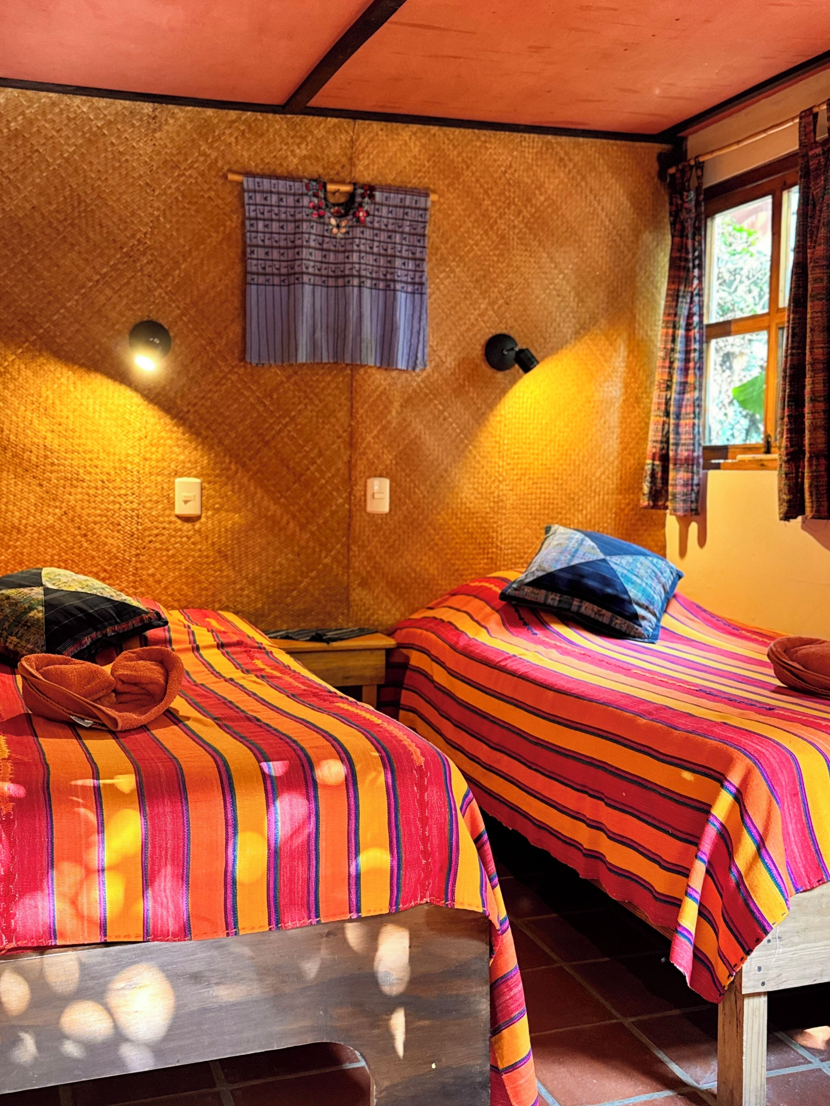 Una habitación con dos camas, cada una con ropa de cama y almohadas a rayas, una mesita, una lámpara de pared y un ventanal con cortinas.