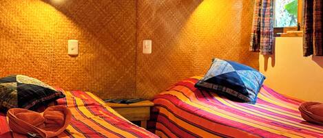 A room with two beds, each with striped bedding and pillows, a small table, a wall-mounted light, and a window with curtains.
