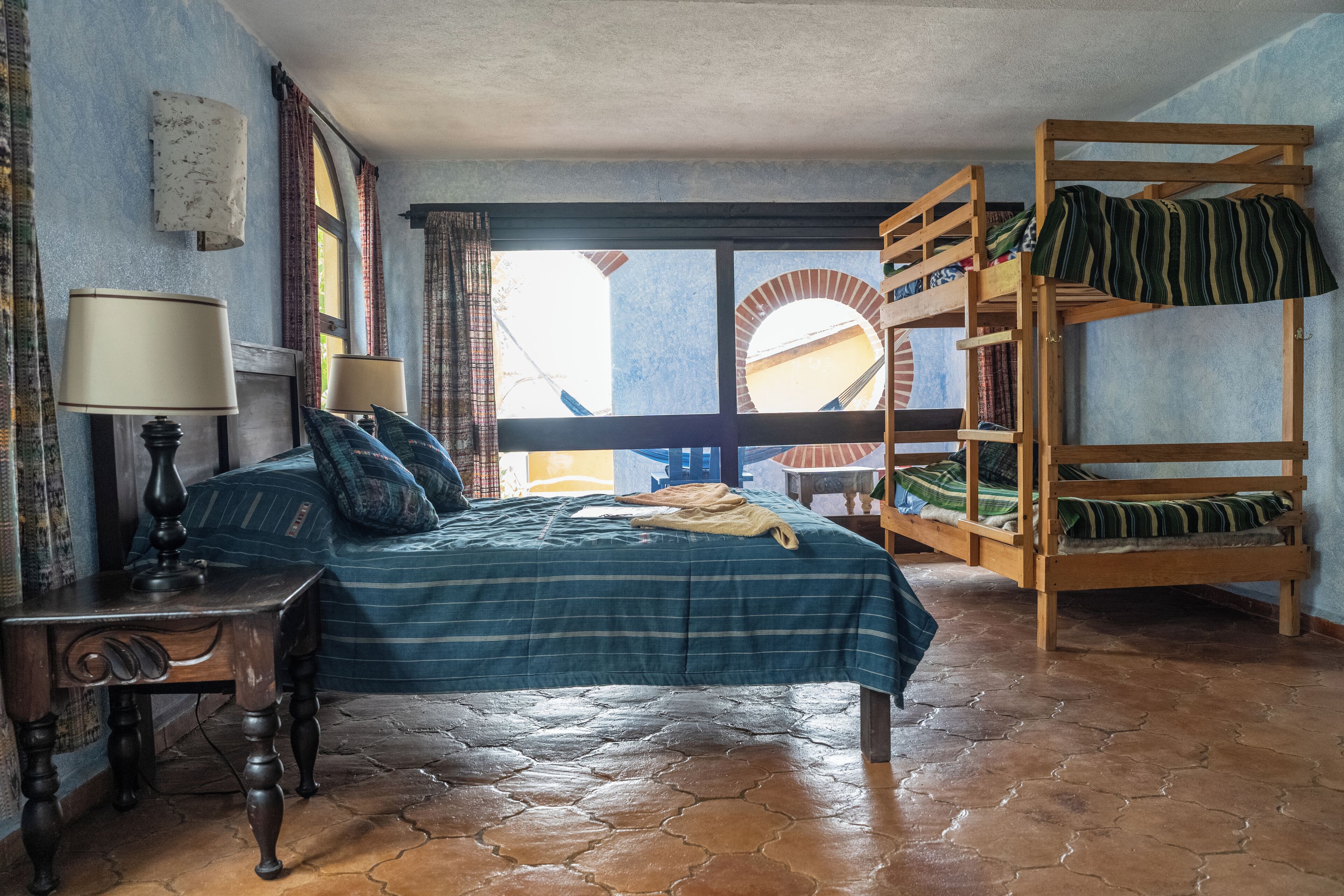 Una habitación con una cama, una litera de madera, una mesa con lámparas y vistas por una ventana.