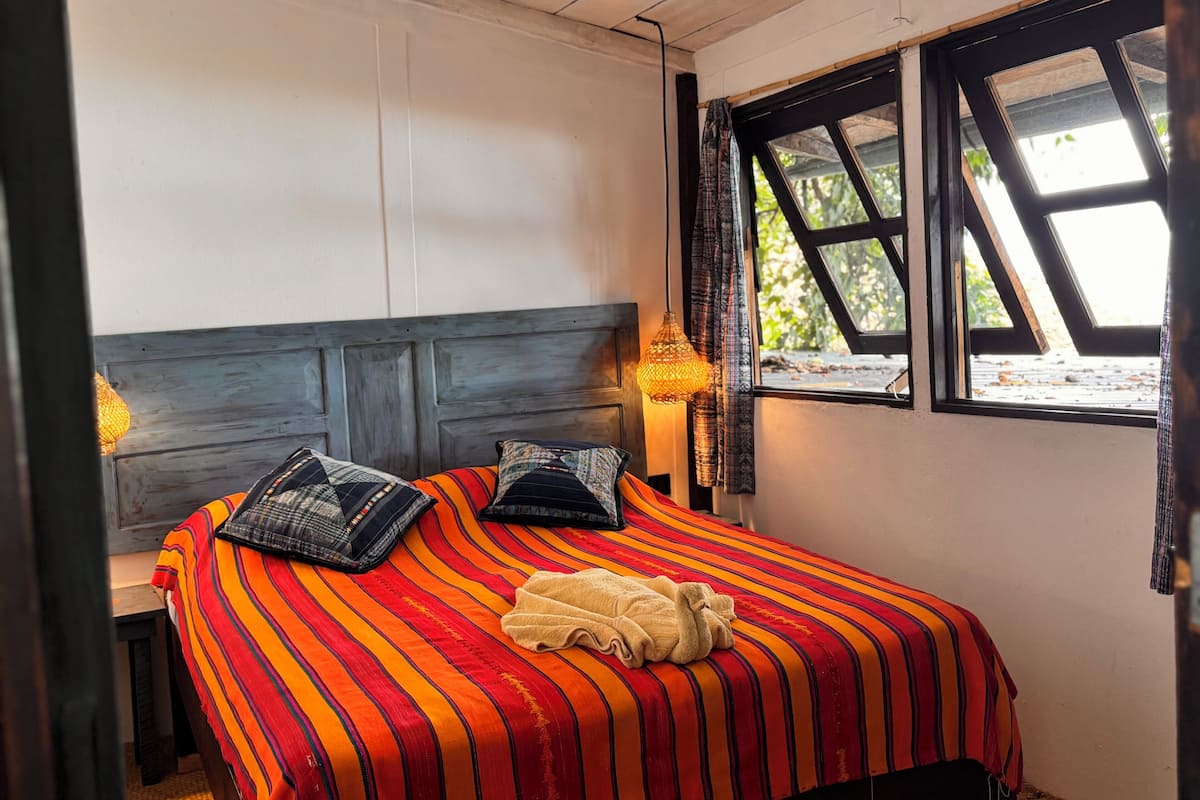 Ein Bett mit gestreifter Bettdecke, zwei Kissen, eine Nachttischlampe und ein Fenster mit Blick auf Grün.
