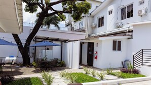 Um pátio com uma mesa e cadeiras sob um guarda-sol azul, cercado por plantas e um edifício branco com janelas e aparelhos de ar condicionado.