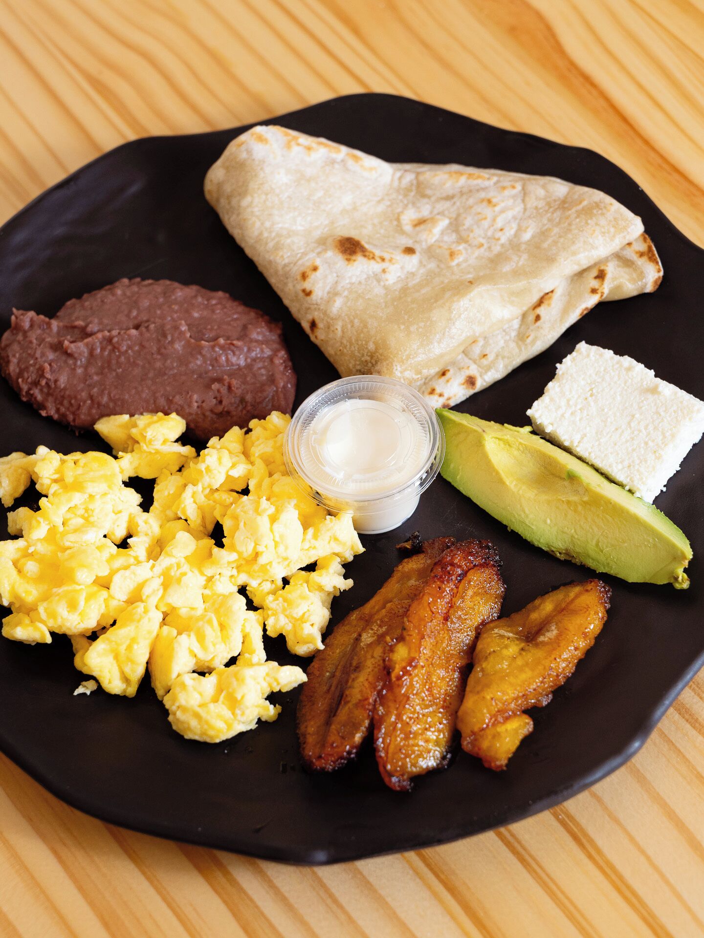 Une assiette contenant des œufs brouillés, des haricots rouges en purée, de l’avocat, des bananes plantains frites, une tortilla et un petit pot de crème aigre.
