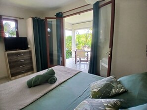 Een slaapkamer met een bed, een televisie op een kast, en uitzicht op een terras met stoelen en begroeiing.