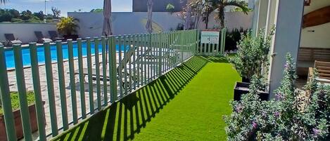A pool area with a green artificial turf path, palm trees, and a white fence.