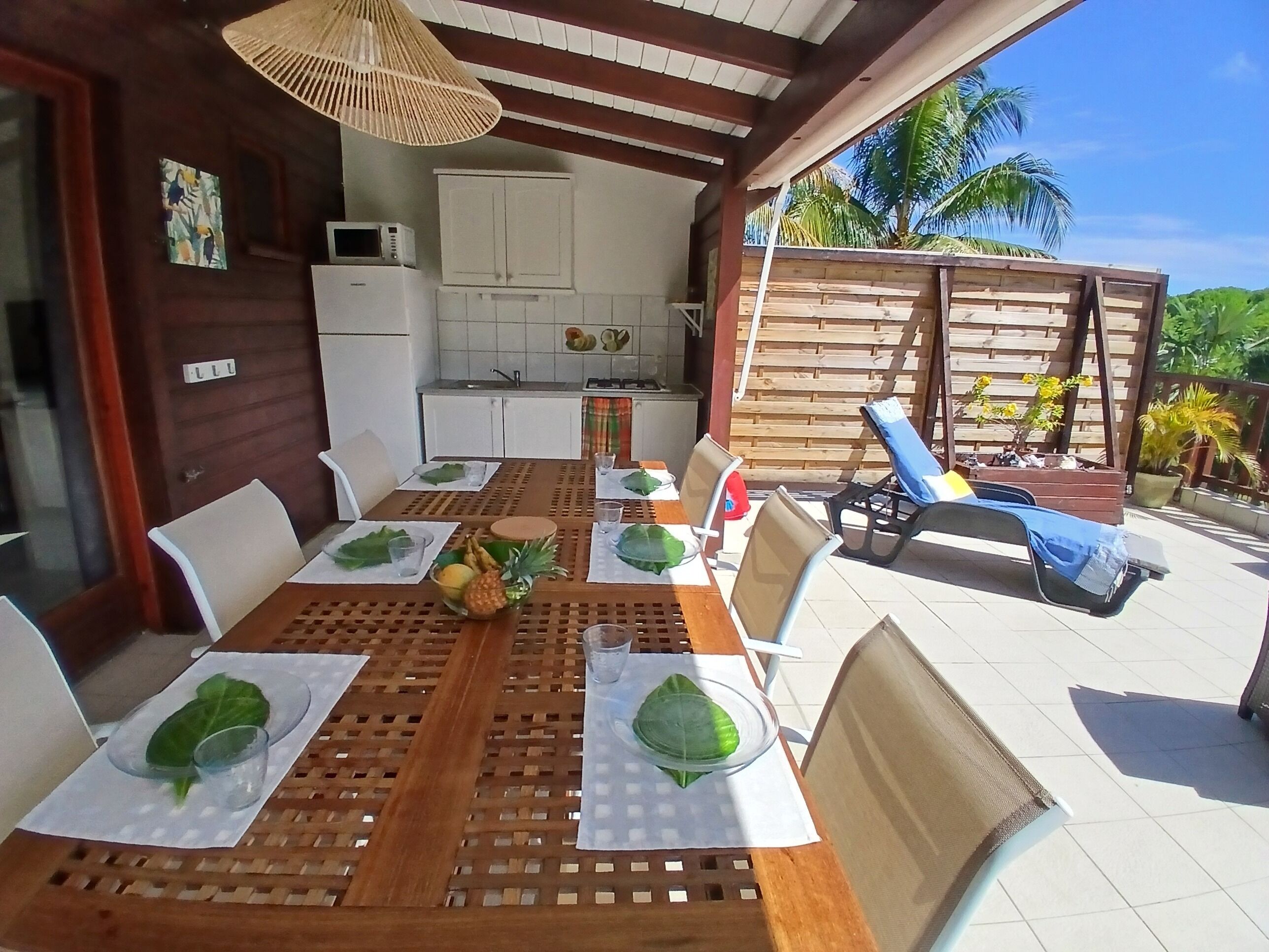 A wooden dining table with chairs, a woven ceiling light, a kitchen area, and a wooden fence with plants.