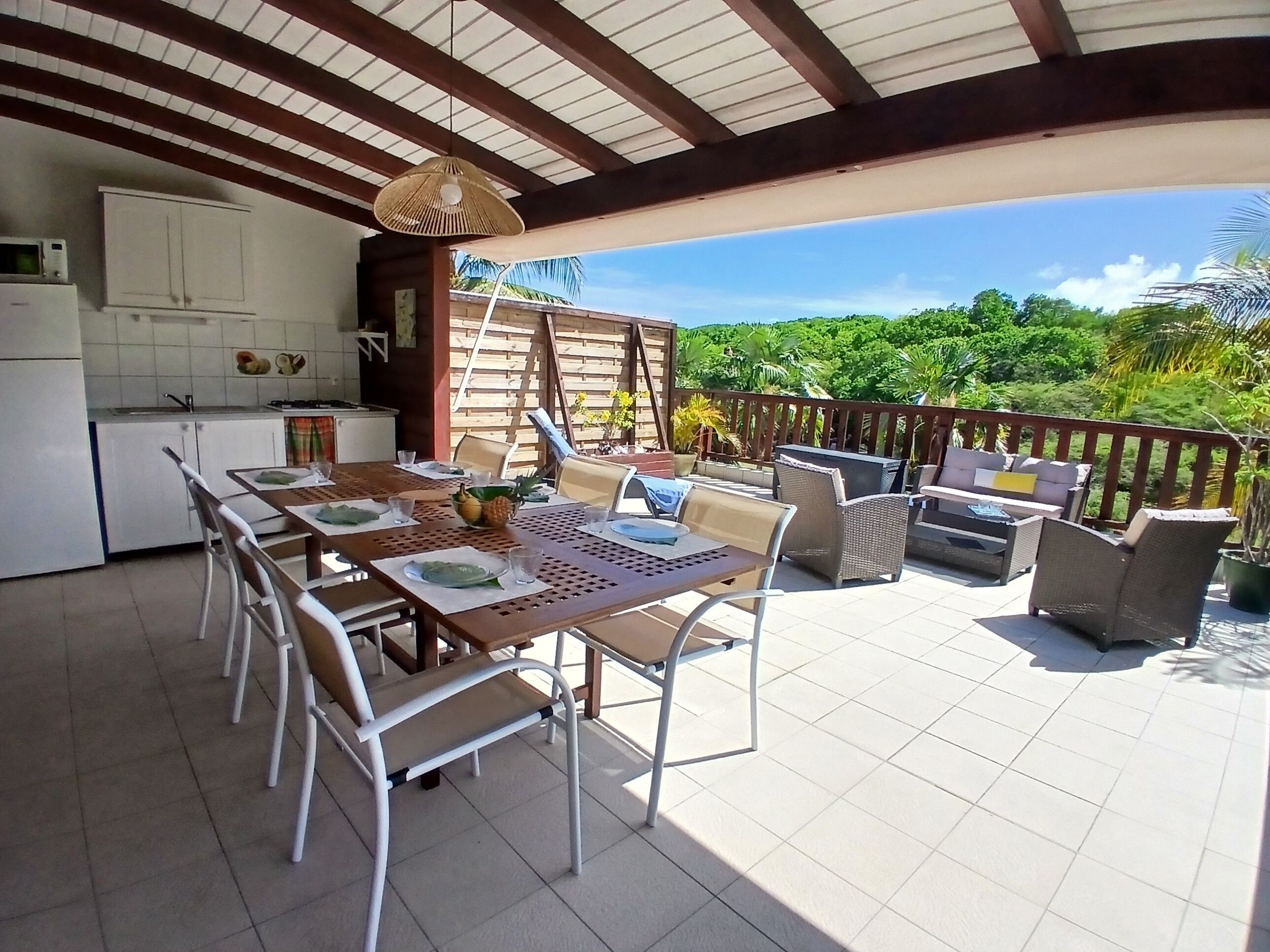 En overdækket terrasse med spisebord og stole, et tekøkken samt udsigt til frodig vegetation.