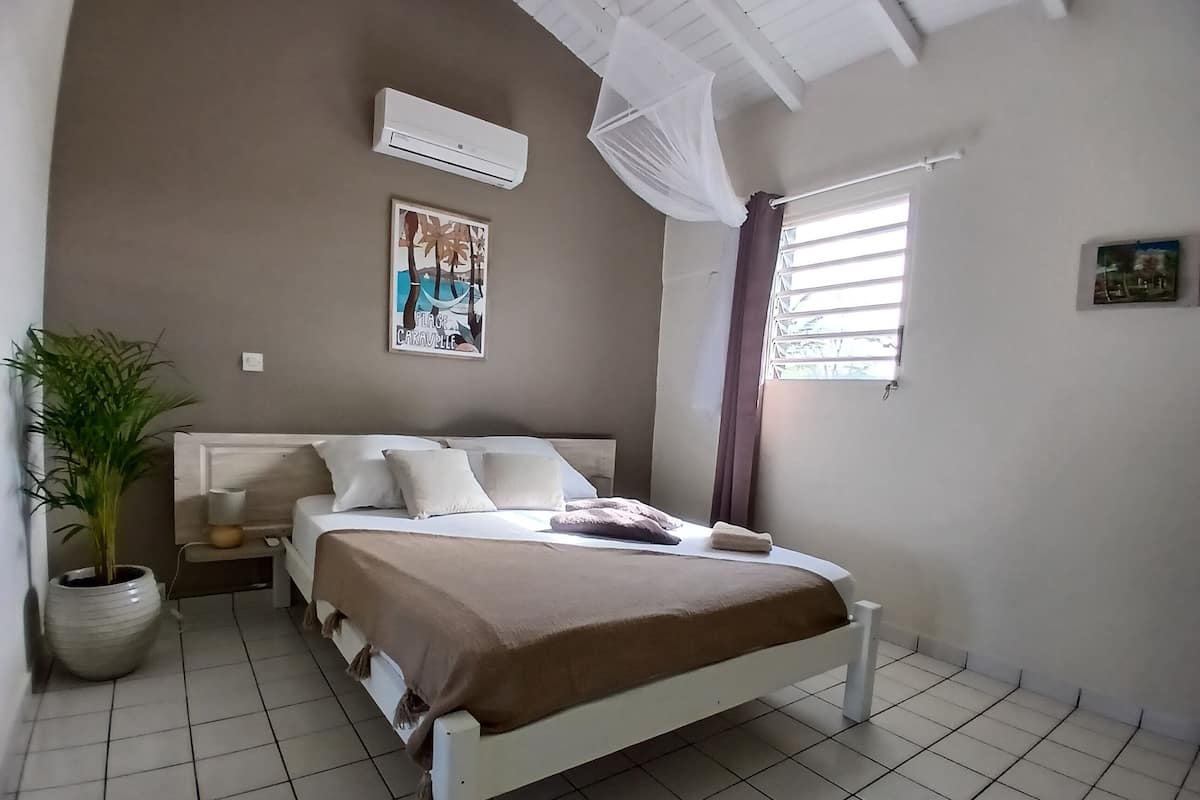 A bedroom with a bed, a potted plant, a wall-mounted air conditioner, and a window with curtains.