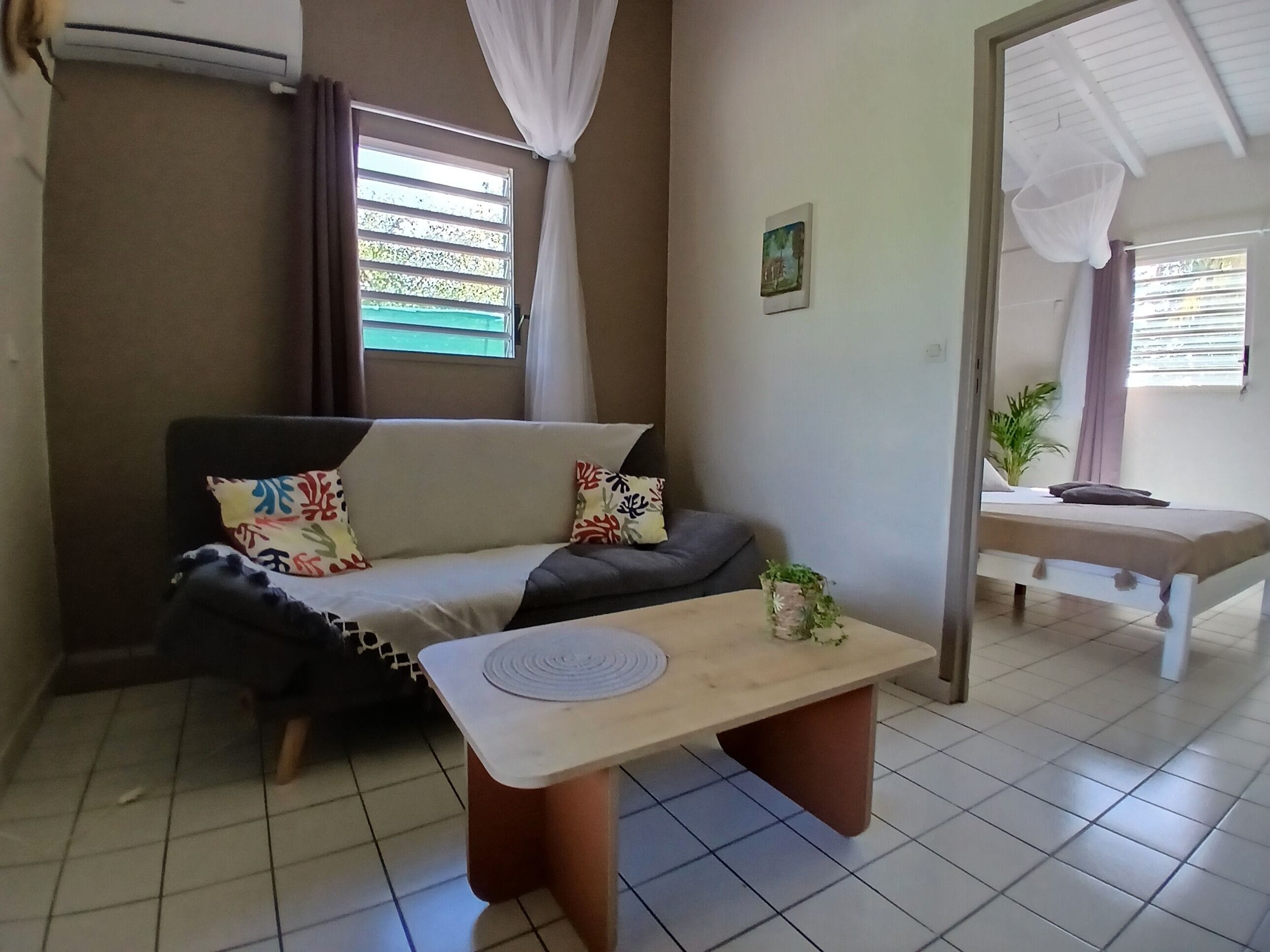 A room with a sofa, a small table, a bed, a window with curtains, and a plant.