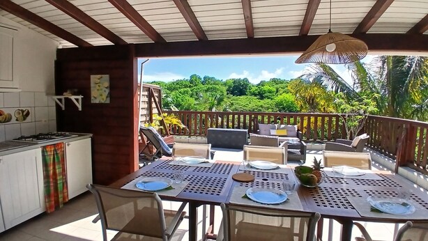 A covered outdoor dining area with a table set for four, a kitchen area with a gas stove, and a view of lush greenery. - Residence Ti Village Créole (Sainte-Anne)