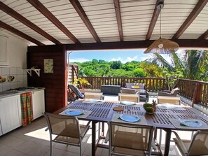 A covered outdoor dining area with a table set for four, a kitchen area with a gas stove, and a view of lush greenery. - Residence Ti Village Créole (Sainte-Anne)
