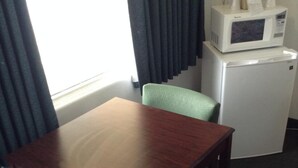 A small wooden table with two green chairs, a white microwave, and a refrigerator in a compact room with a window and curtains. - Plaza Inn Great Falls (Great Falls)