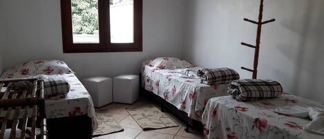 A small, clean room with two single beds, a wooden chair, a small table, and a window with a view of greenery.