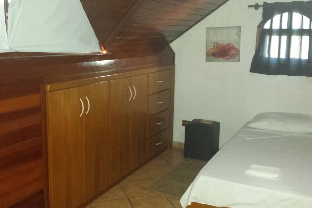 A bedroom with a wooden wardrobe, a bed, a small trash bin, and a window with curtains.