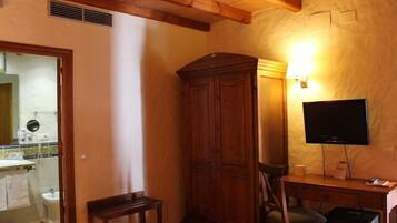 Una habitación con un armario de madera, un escritorio con silla, televisor y un baño visible a través de una puerta abierta.