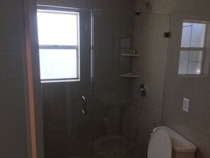 A bathroom with a glass shower enclosure, a toilet, and a window. - Pelican By The Sea (Pompano Beach)