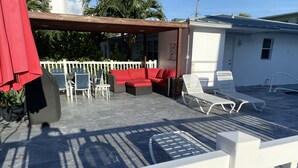 A covered patio area with outdoor furniture, including a red sofa and white lounge chairs. - Pelican By The Sea (Pompano Beach)