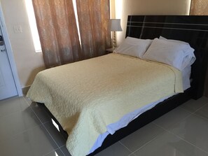 A neatly made bed with a beige bedspread and white pillows in a hotel room with a tiled floor and a patterned curtain. - Pelican By The Sea (Pompano Beach)