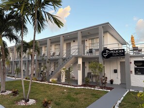 A multi-story hotel with a modern design, featuring a balcony on the second floor, a sign reading "PELICAN By The Sea," and a well-maintained garden with palm trees and shrubs. - Pelican By The Sea (Pompano Beach)
