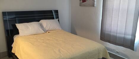 A bed with a headboard, white pillows, and a beige bedspread in a room with a window and a wall-mounted picture.