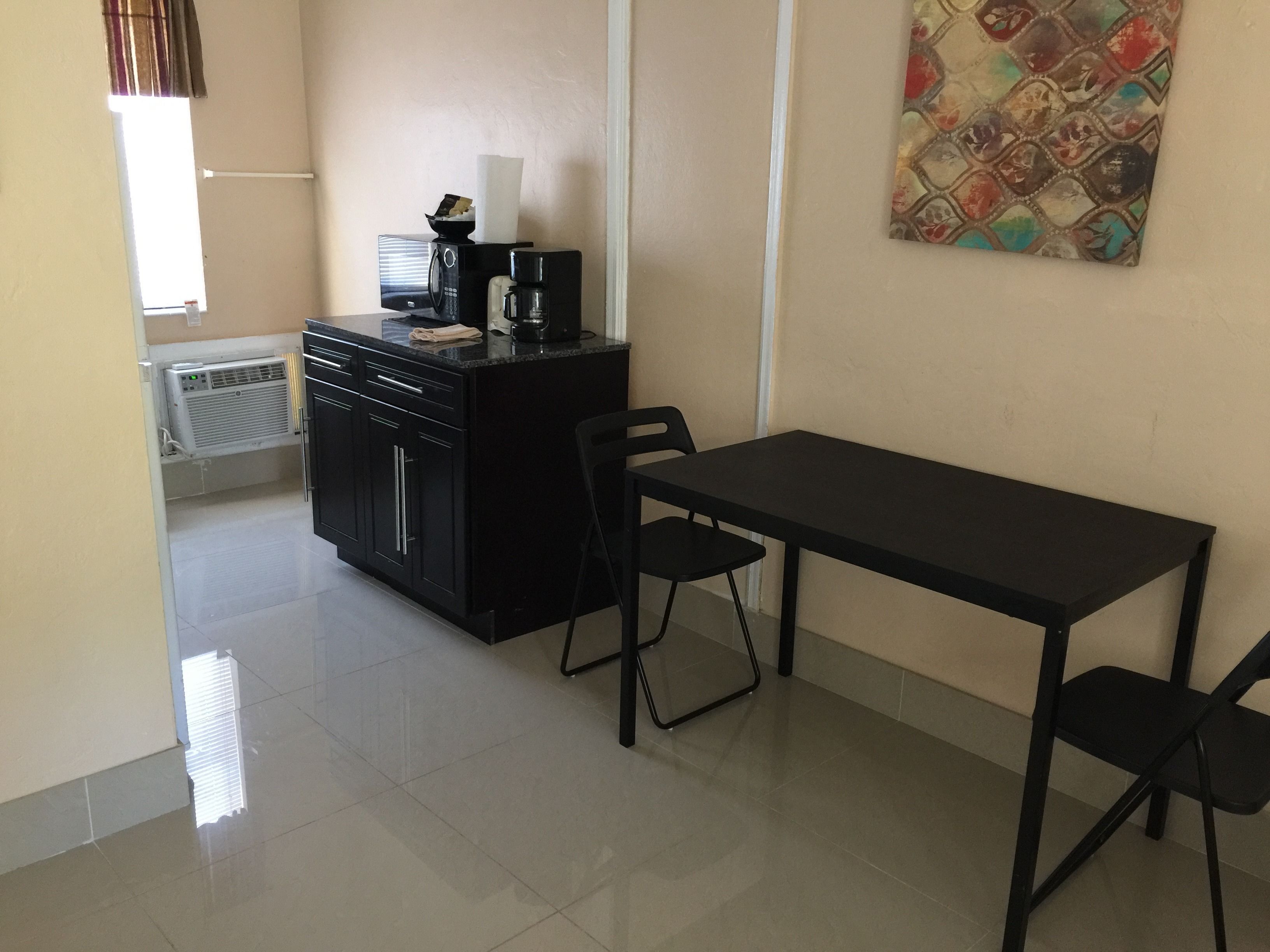 A small kitchenette with a coffee maker, a microwave, and a refrigerator. There is a black table with two chairs and a painting on the wall.