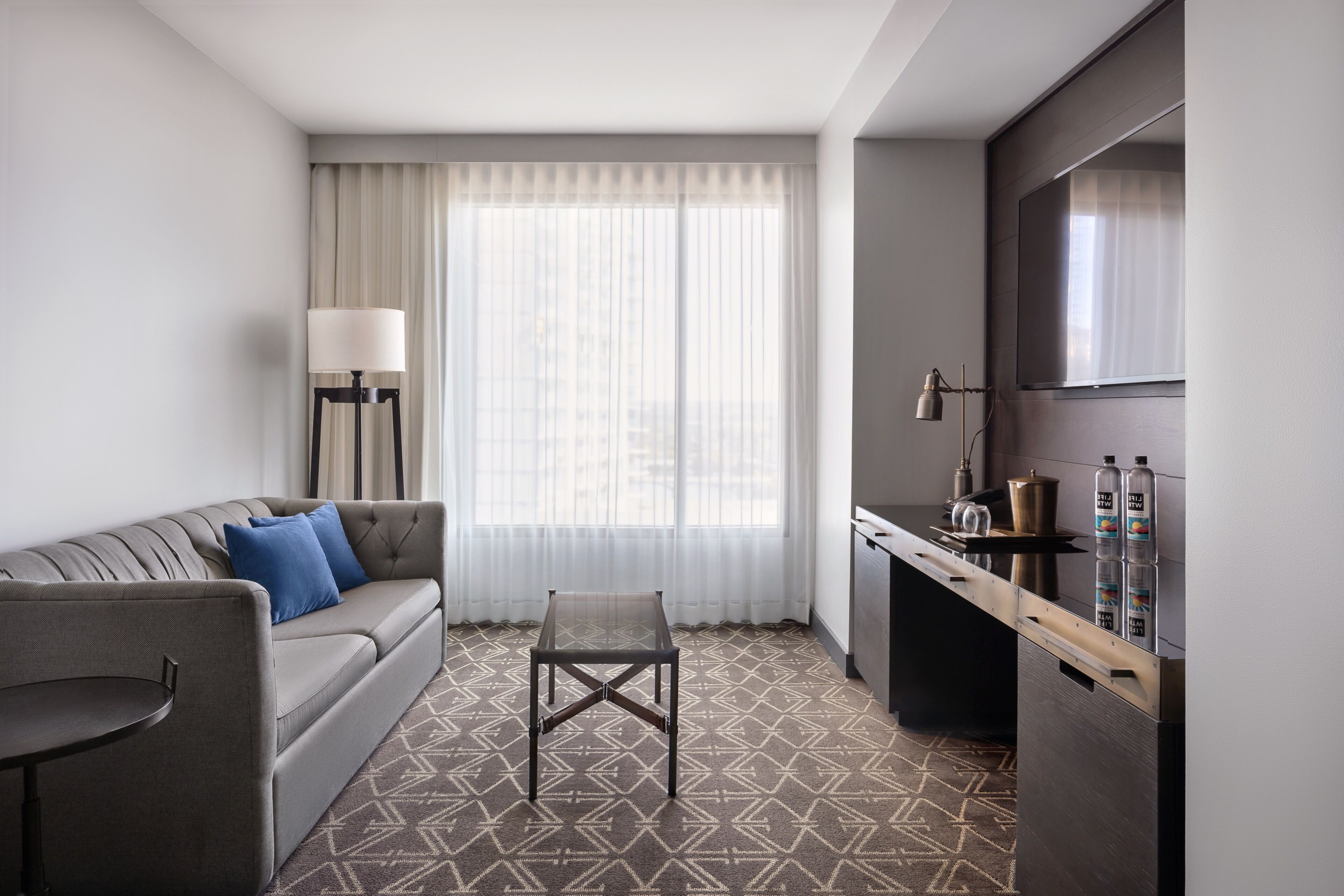 A modern hotel room with a sofa, a small table, a desk, and a large window with curtains.