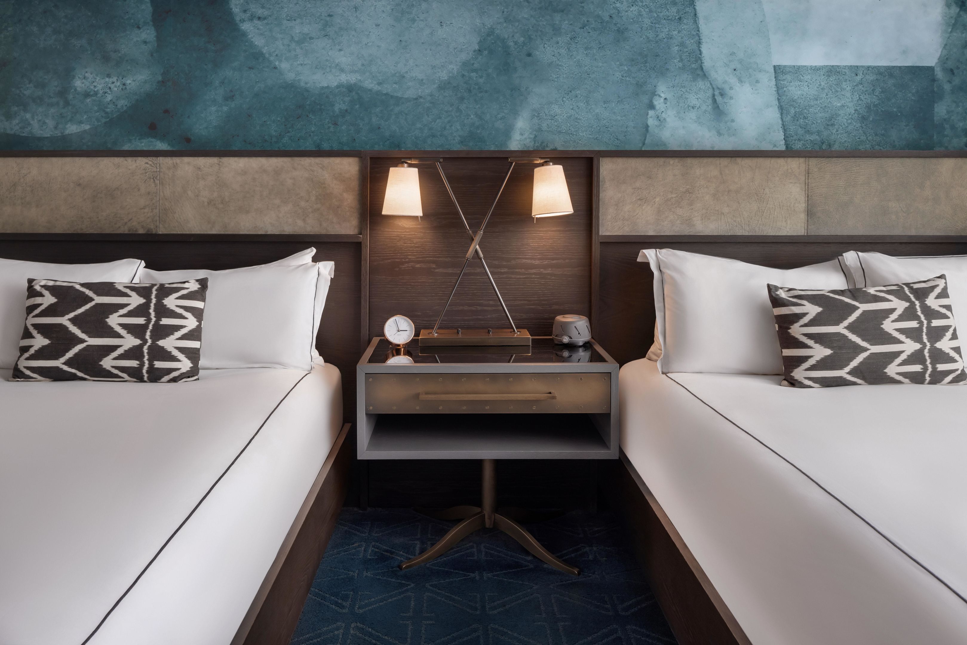 A hotel room with two beds, a nightstand with clocks, and a wall-mounted light fixture.