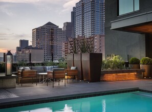 Un área de piscina en la azotea con mobiliario, un jardín con plantas y un horizonte urbano al fondo.