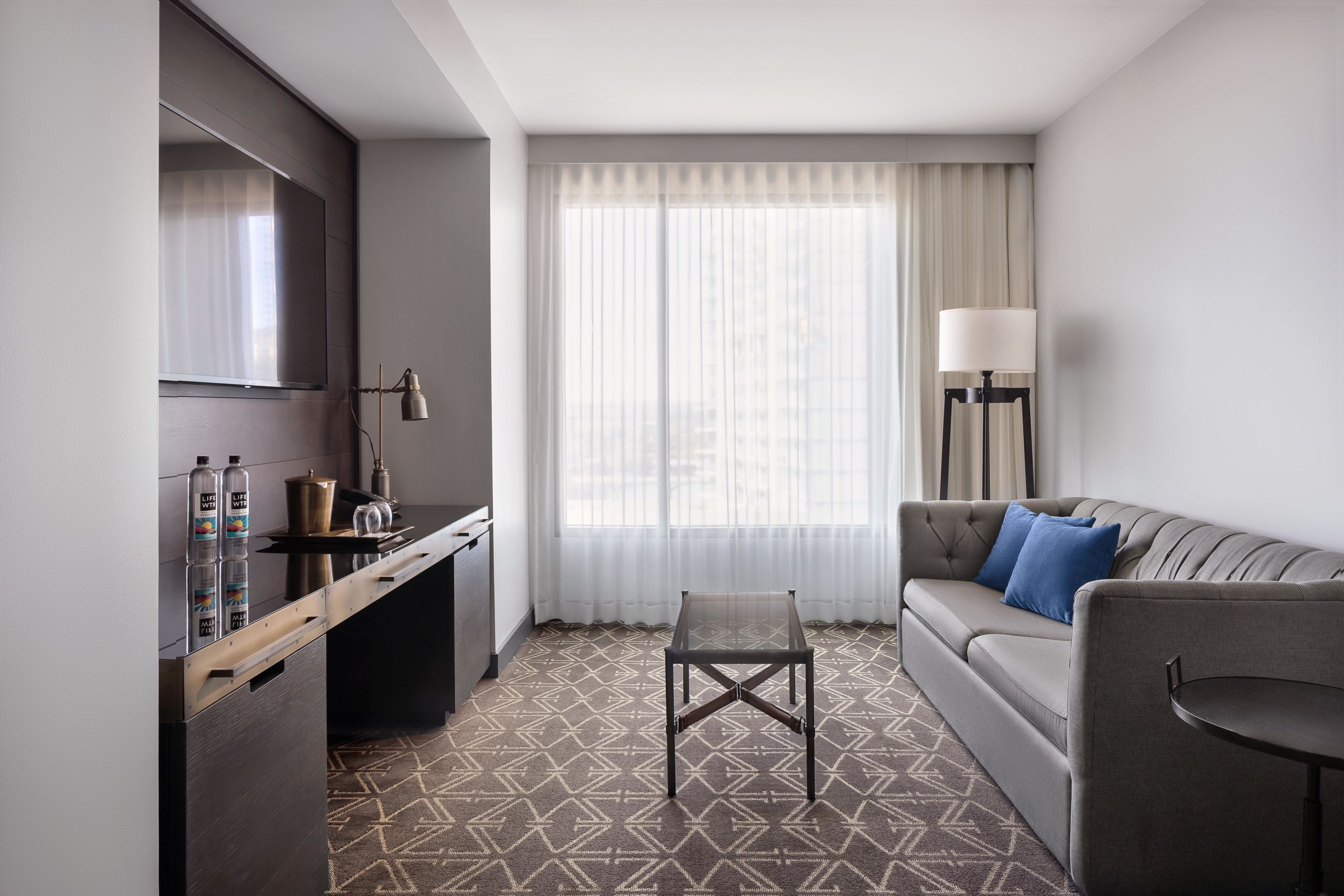 Une chambre d’hôtel équipée d’une kitchenette, d’un canapé, d’une table basse et d’une grande fenêtre avec des rideaux légers.