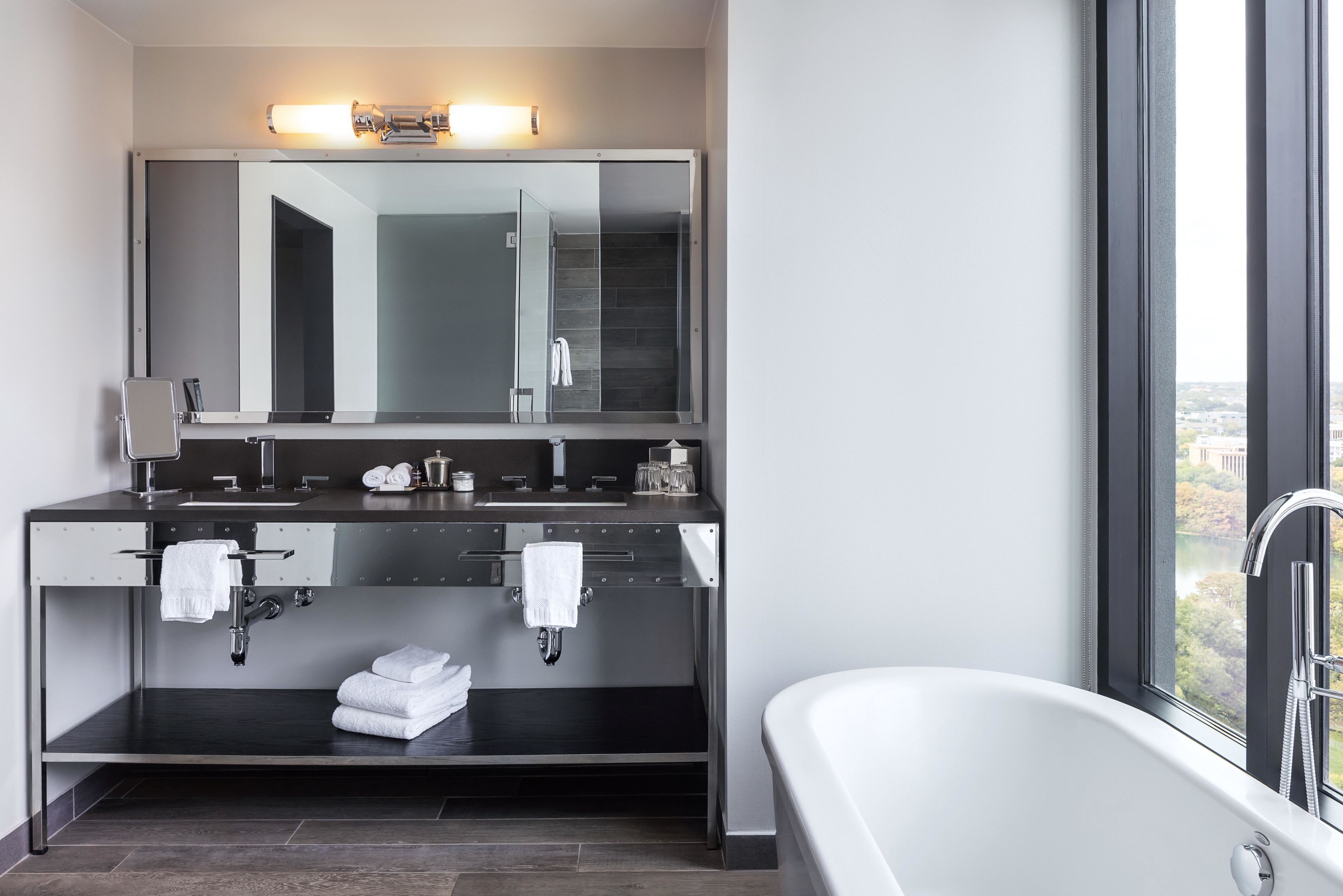 A modern bathroom with a large mirror, dual sinks, a bathtub, and a view of the city through a large window.
